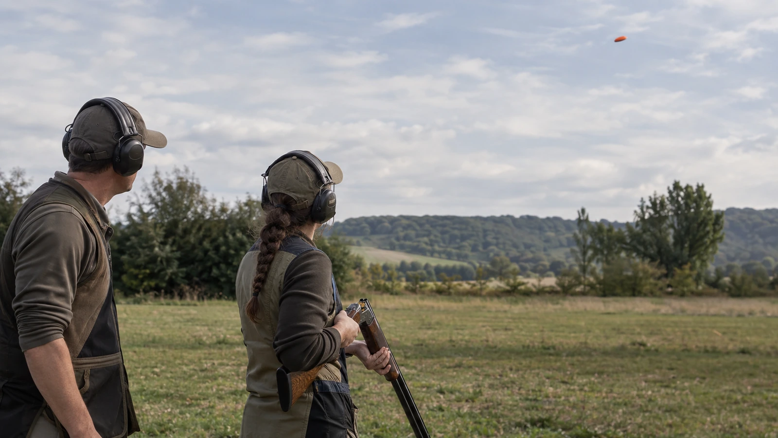 Deux personnes portant des protections auditives sur un terrain de ball-trap ; la tireuse tient son fusil ouvert et orienté vers le sol